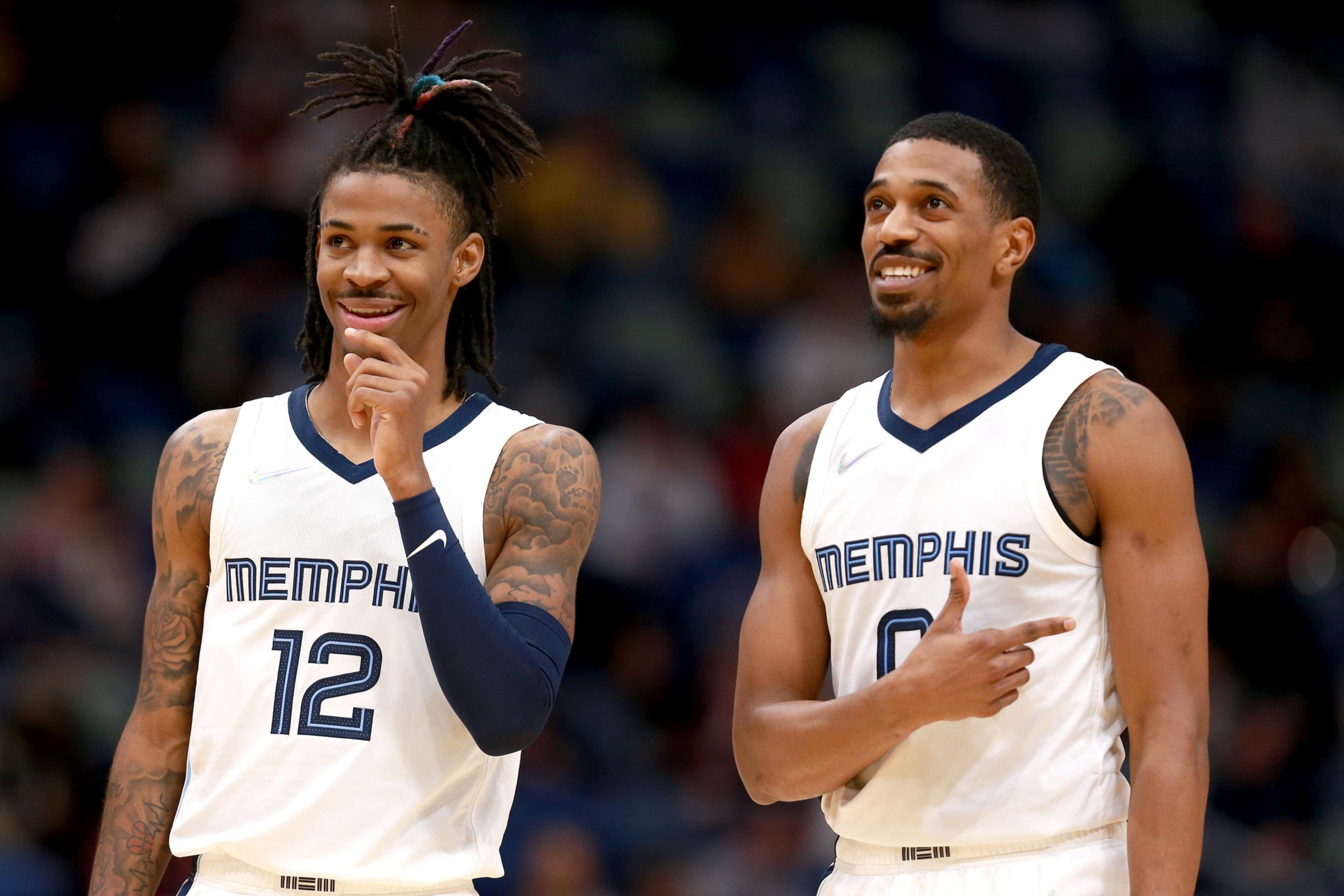 Two basketball players in white Memphis jerseys, numbers 12 and 0, smile and gesture while standing on the court during a game.