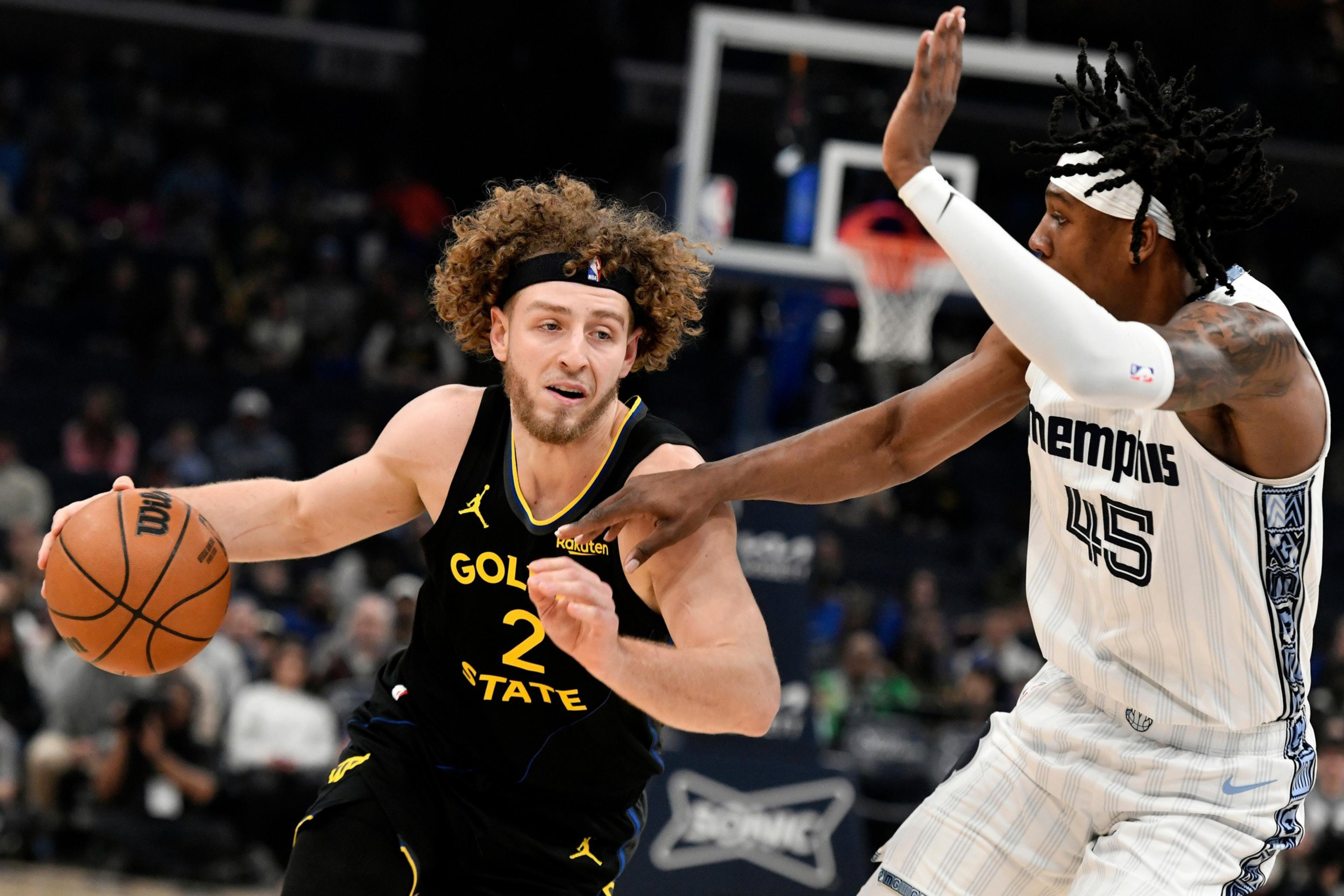A Golden State player with curly hair drives past a Memphis defender wearing white, both focused intensely as they compete near the basket.