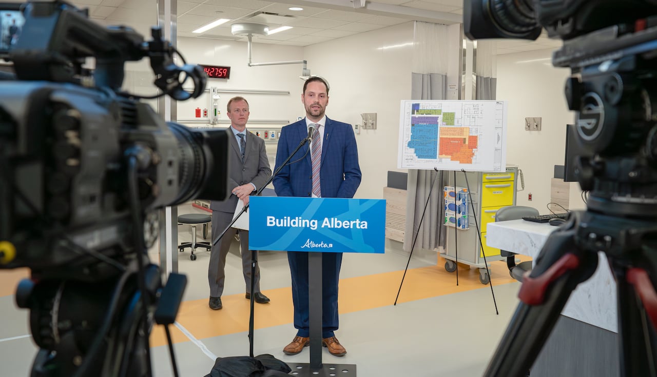 A man stands at a podium in a hospital with cameras pointing at him. Another man is standing behind him.