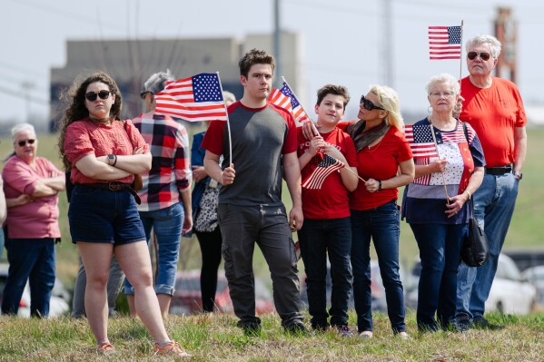 People holding U.S. flags await the hearse and procession for Army Sgt. Benjamin N. Pennington, 26, of Glendale, Ky, March 20, 2026, in Elizabethtown, Ky. (AP Photo/Jon Cherry)