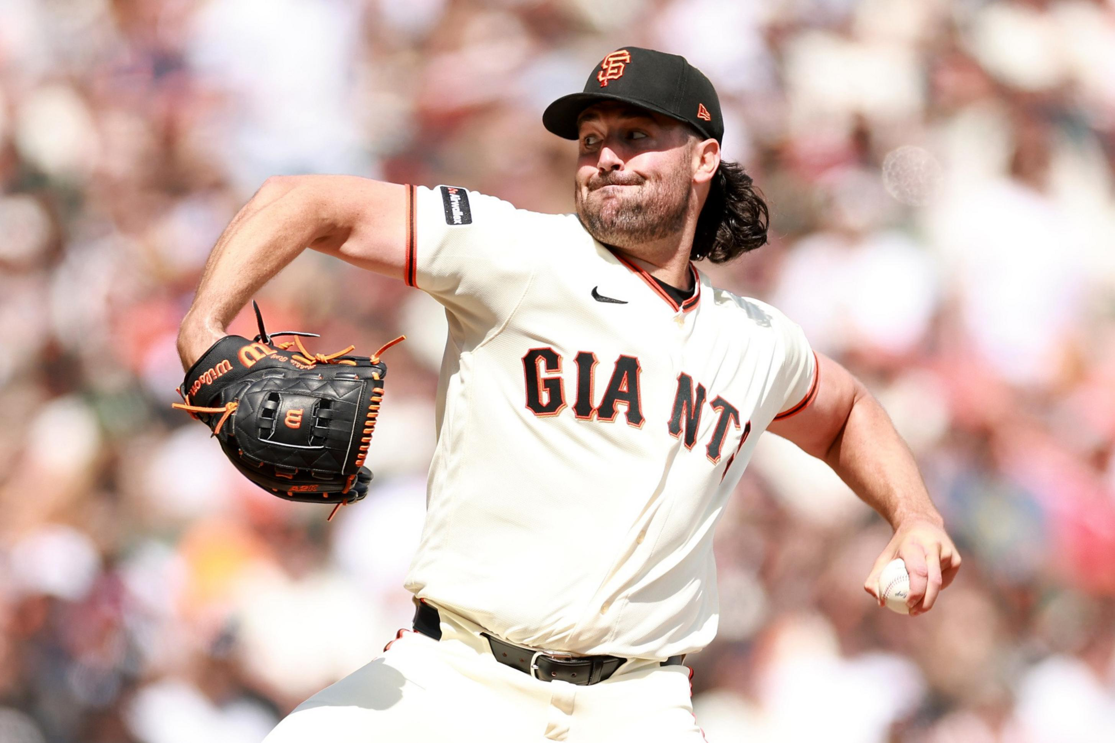 A baseball pitcher wearing a San Francisco Giants uniform is in mid-throw with a focused expression, gripping a baseball in his right hand.