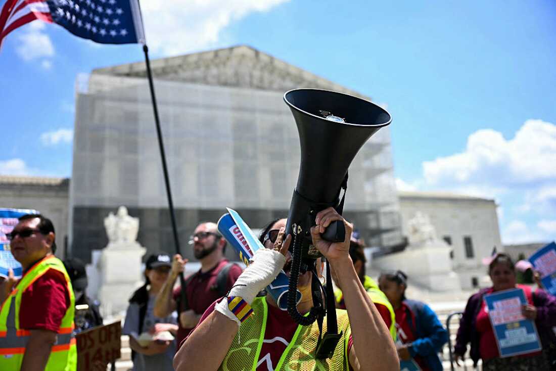 Protesters stand outside the U.S. Supreme Court. One holds a flag, another has a bullhorn, and some hold signs.