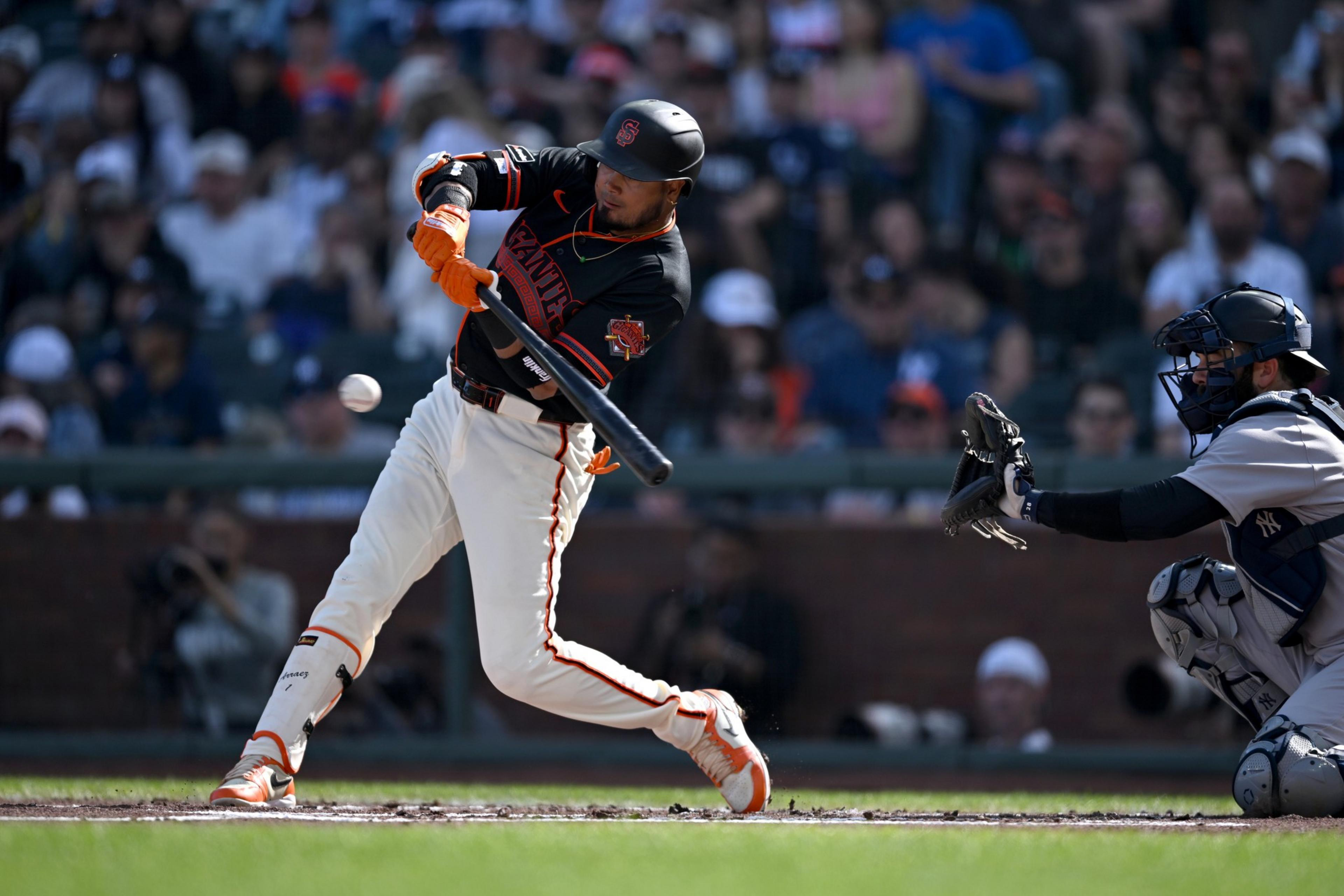 A baseball batter in a black and orange uniform swings at a pitch while a catcher in gray and navy gear prepares to catch the ball.