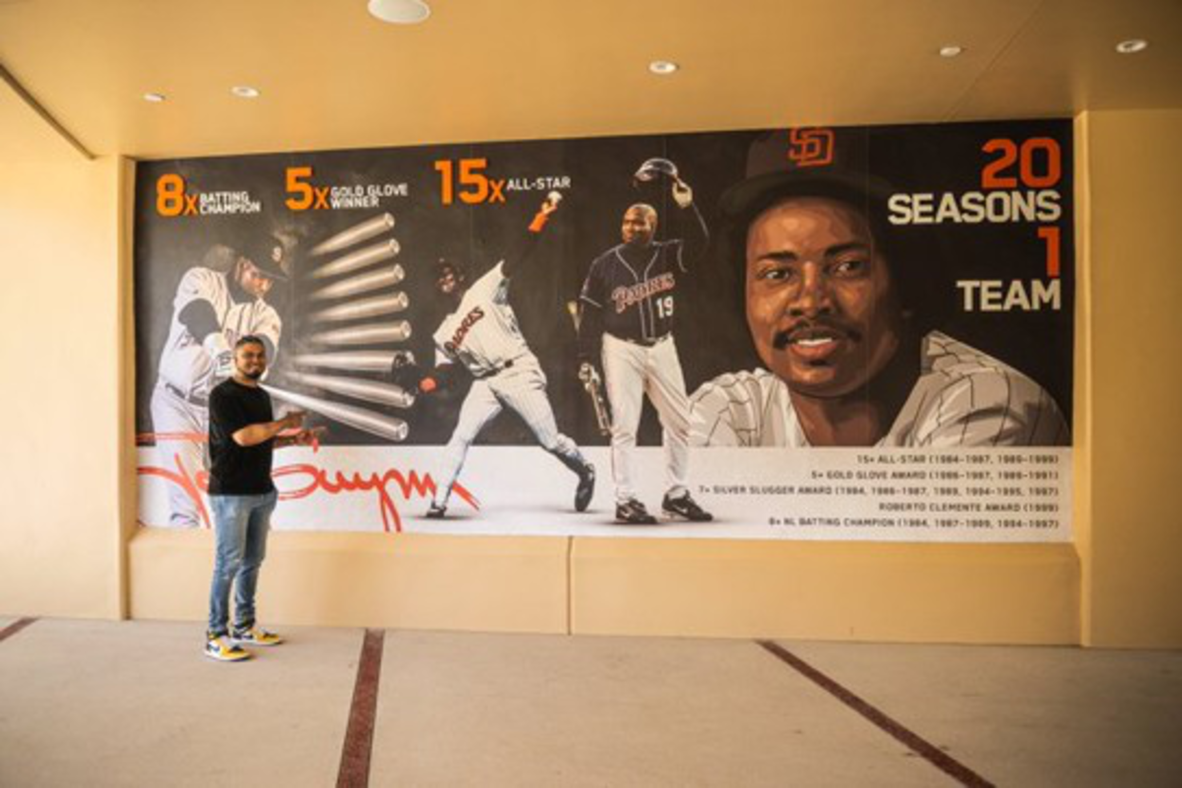 A man stands beside a large mural celebrating a baseball player’s 20 seasons, 8 batting titles, 5 Gold Gloves, and 15 All-Star selections.