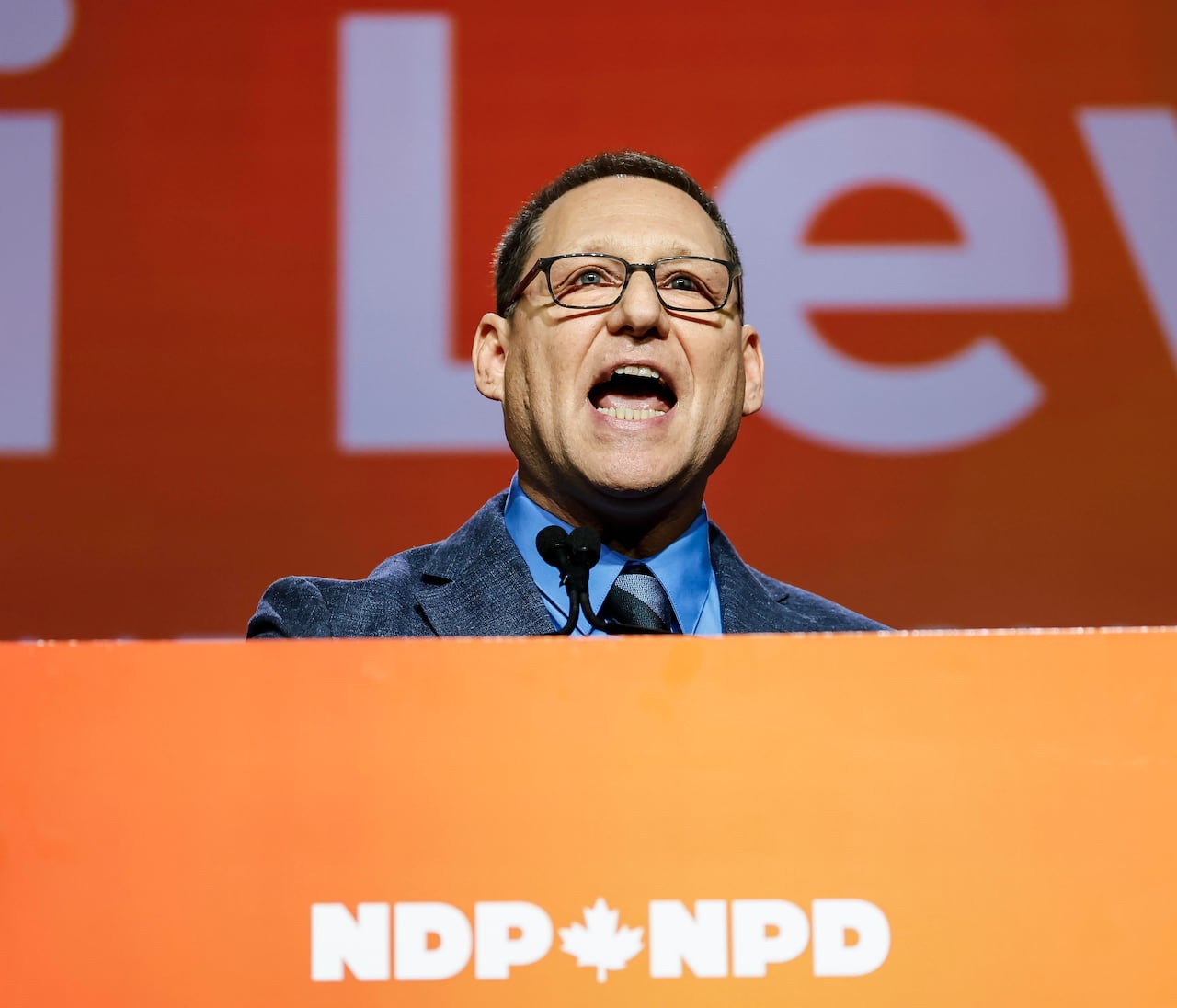 A man in a suit stands at an orange podium.