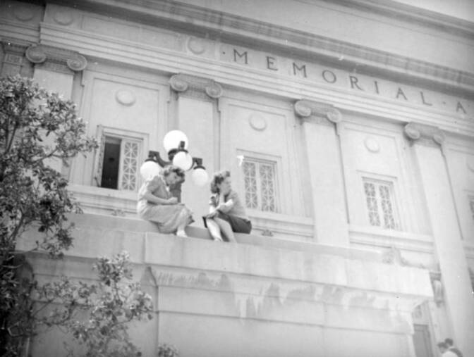 A black and white photo of a Roman-temple style building. Two women sit on a concrete ledge.