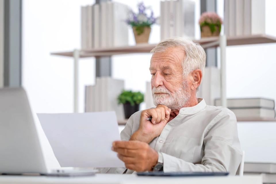 A senior man with white hair and beard, wearing a light-colored collared shirt, sits at a desk. He holds a white paper document in his left hand and a pen in his right, resting his chin as he looks intently at the document, suggesting careful thought. A silver laptop is visible to his left, and blurred shelves with books and plants are in the background.