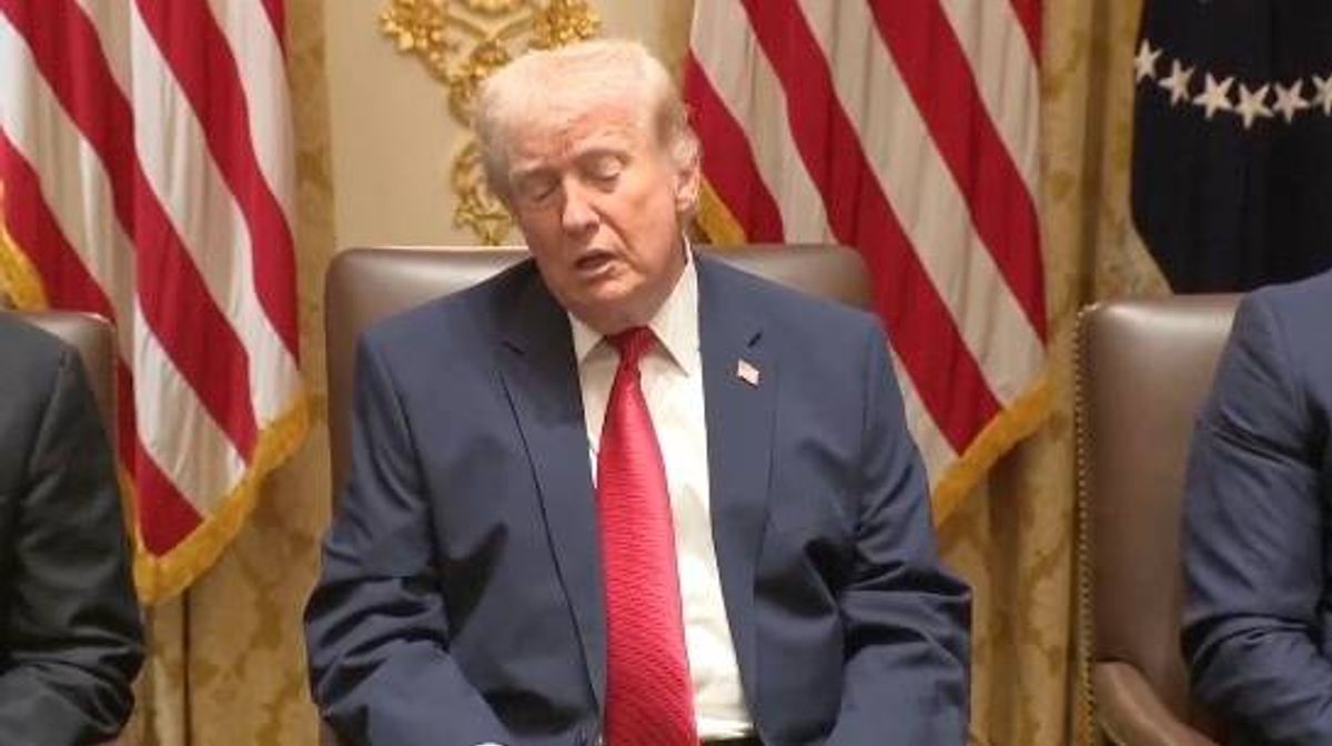 An individual dressed in formal attire, seated in an official setting, possibly engaged in a formal discussion or meeting. The backdrop features American flags, suggesting a context related to political or governmental activities.