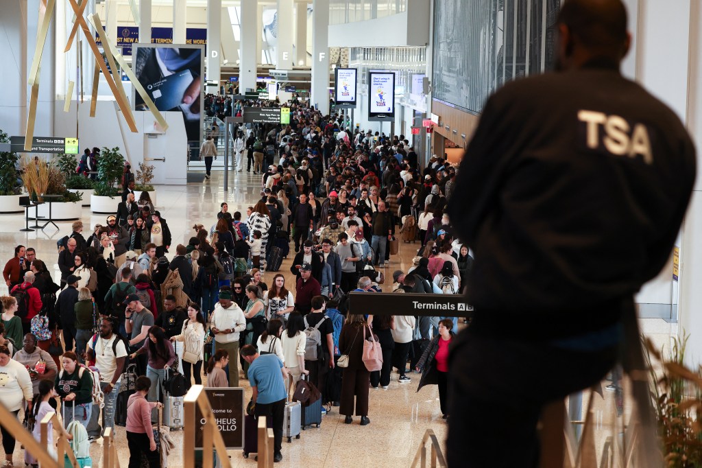 A large crowd of passengers queues at airport security, with a TSA agent in the foreground facing away from the camera.