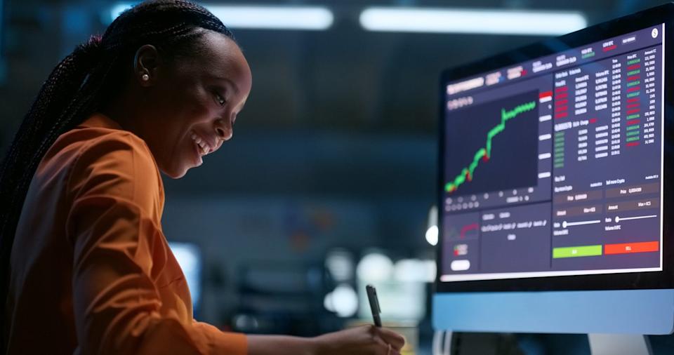 Person smiling and writing while sitting in front of a screen with a stock chart.