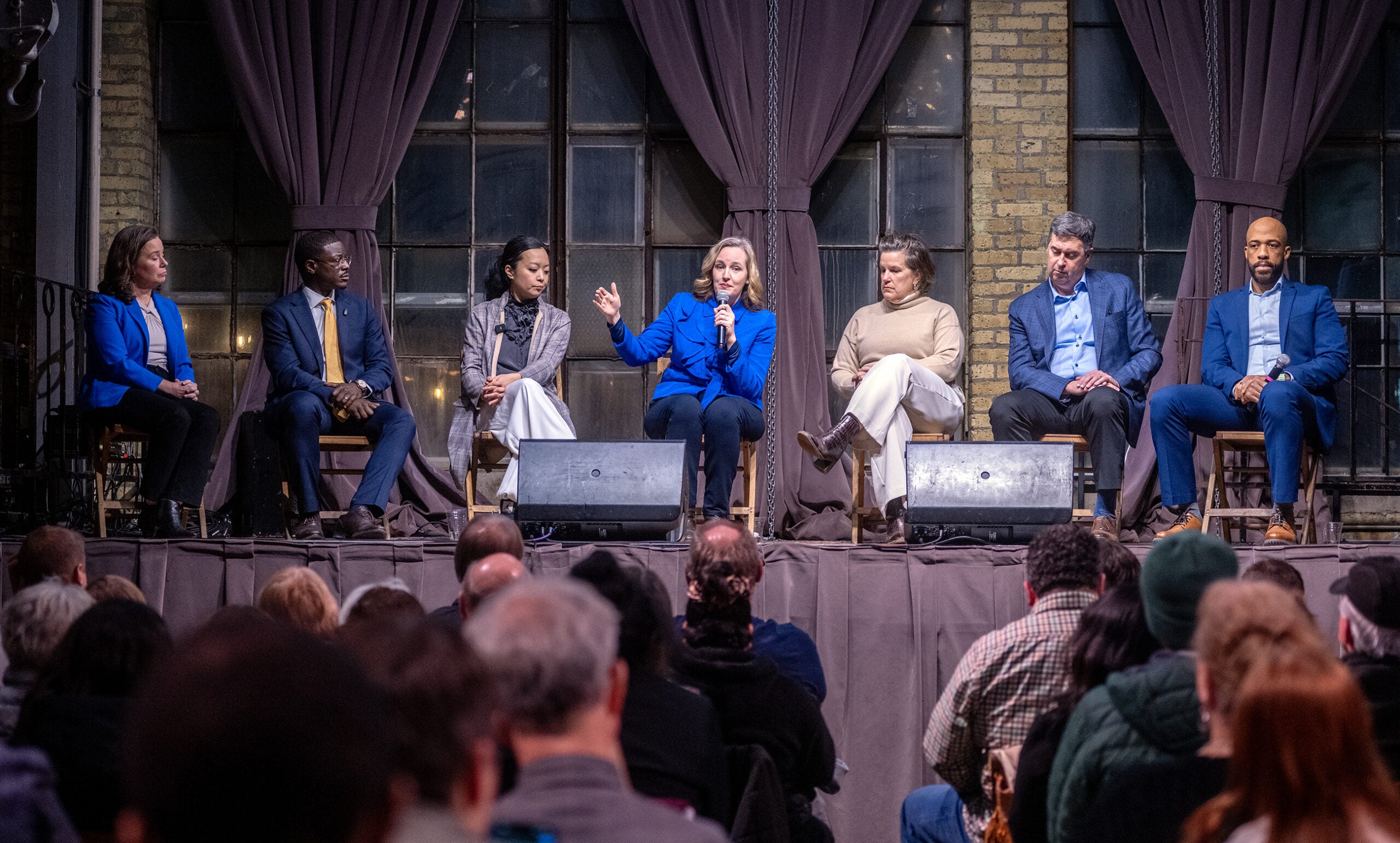 Seven people sit on a stage panel in front of an audience; one person in the center speaks while others listen. The setting appears to be an indoor event with large windows behind them.