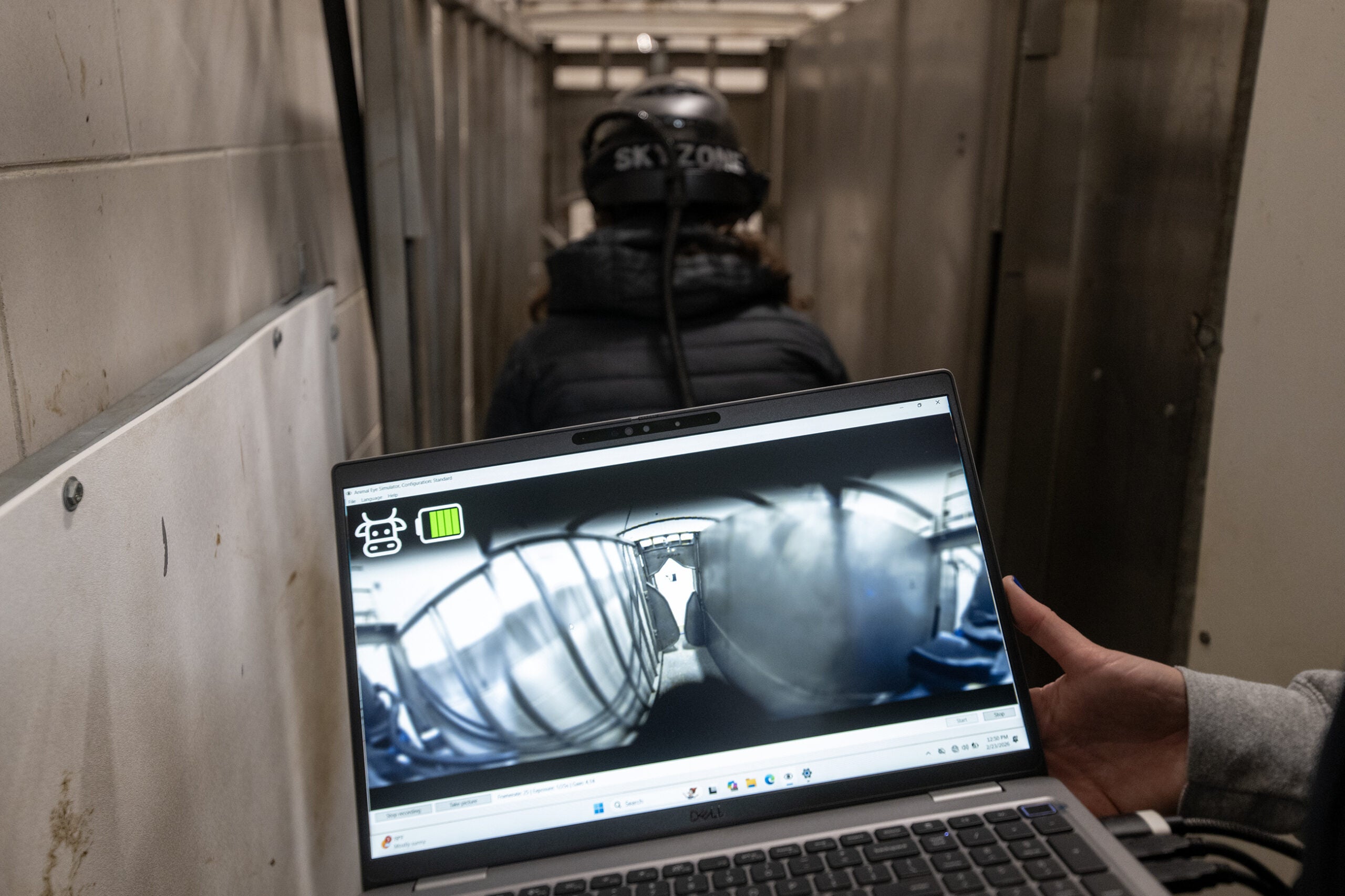 A person holds a laptop displaying a fisheye camera view of a narrow hallway, while another person in a helmet walks ahead in the corridor.