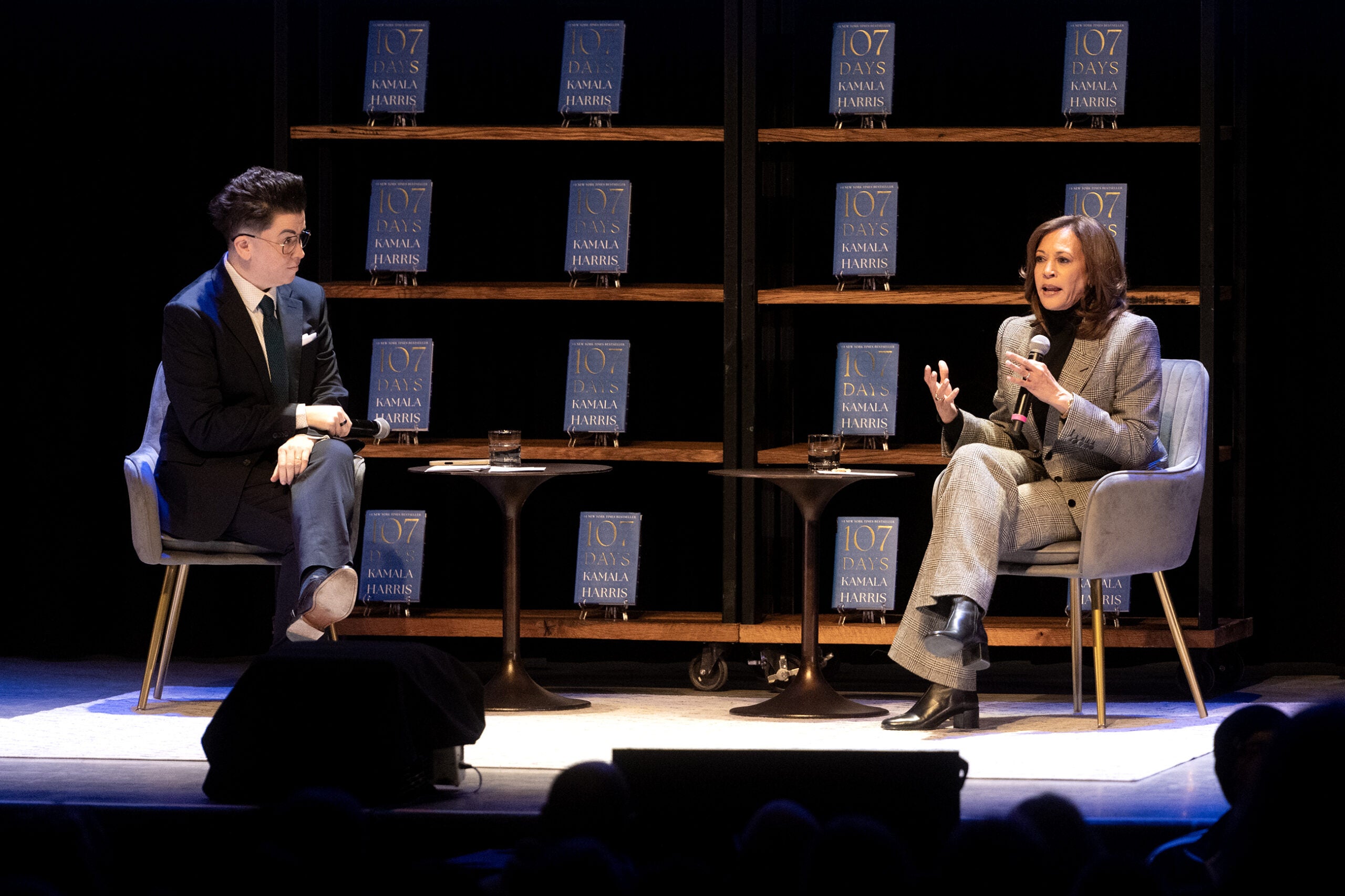 Two people sit and converse on a stage with microphones, surrounded by shelves displaying multiple copies of a book titled 107 Days.