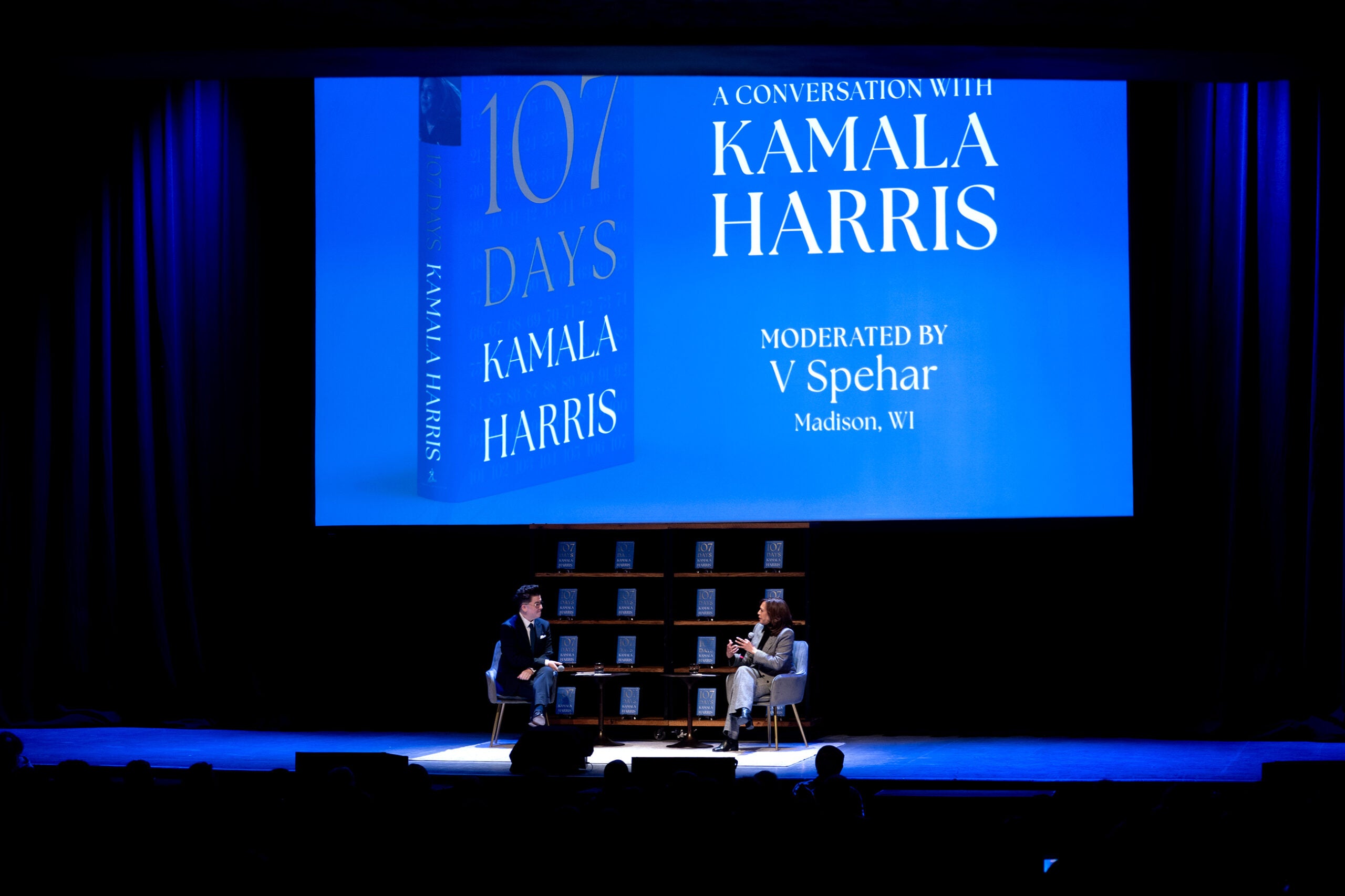 A stage event features Kamala Harris in conversation with a moderator, with a large screen displaying 107 Days and event details in the background.