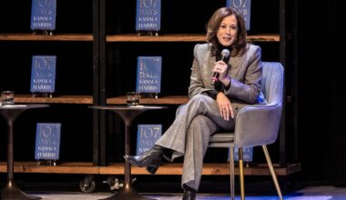 A woman in a gray suit sits in a chair on stage, holding a microphone. Multiple copies of a book titled 107 Days by Kamala Harris are displayed on shelves behind her.