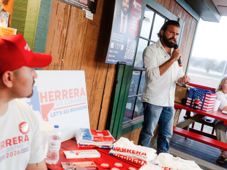 Brandon Herrera speaks into a microphone in front of a small seated crowd, next to a table with his campaign merch