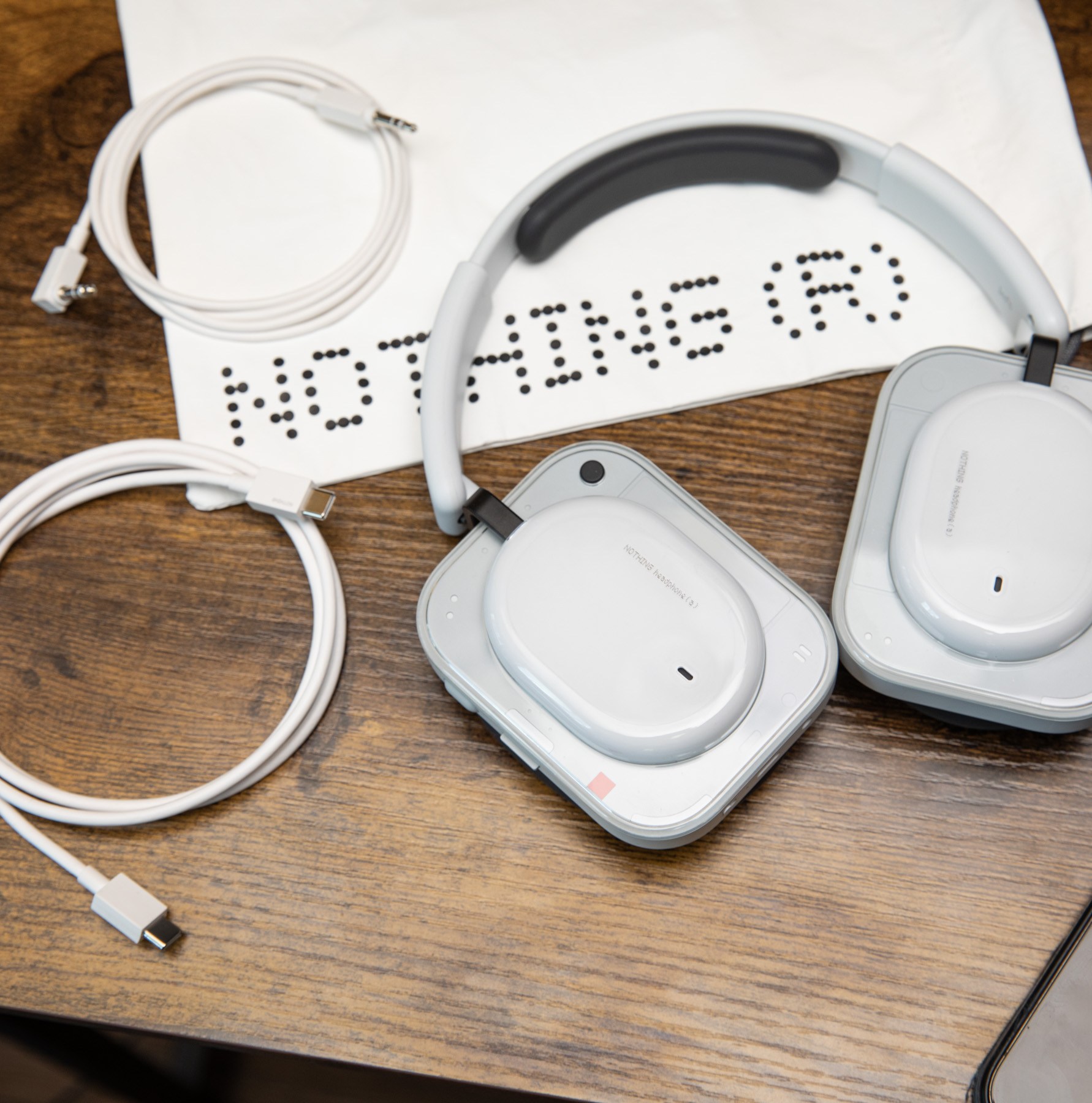 The Nothing Headphone A with its carrying bag, 3.5mm cable, and USB-C cable on a wooden desk with an iPhone nearby.