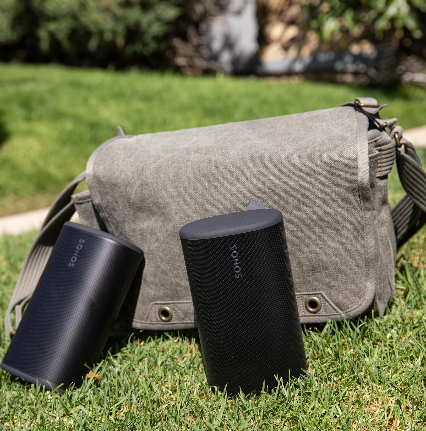Two Sonos Play speakers in front of a sage colored bag on the grass.