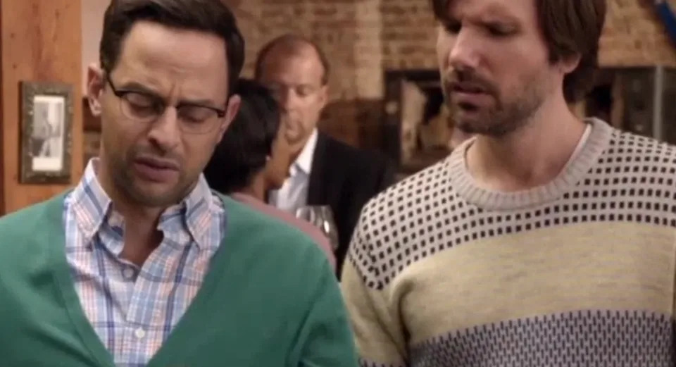 Two men stand together, one in glasses and a cardigan, the other in a striped sweater, appearing puzzled or deep in conversation