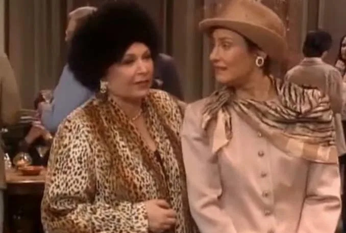 Two women are indoors; one wears a leopard print coat and fur hat, the other wears a beige suit with a hat, scarf, and large earrings