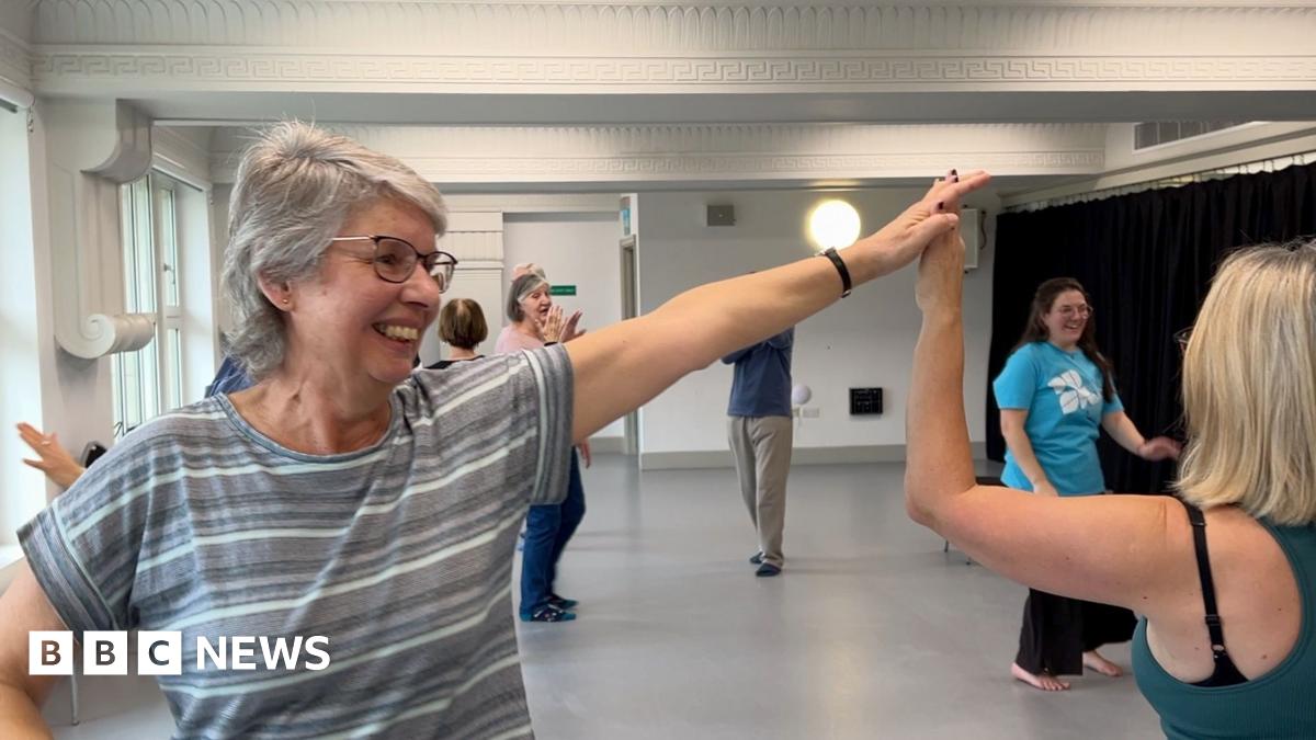 A woman with grey shoulder length hair and a striped top has her left arm outstretched and raised dancing with her partner, whose face isn't visible. A handful of other dancers and an instructor is visible in the background.