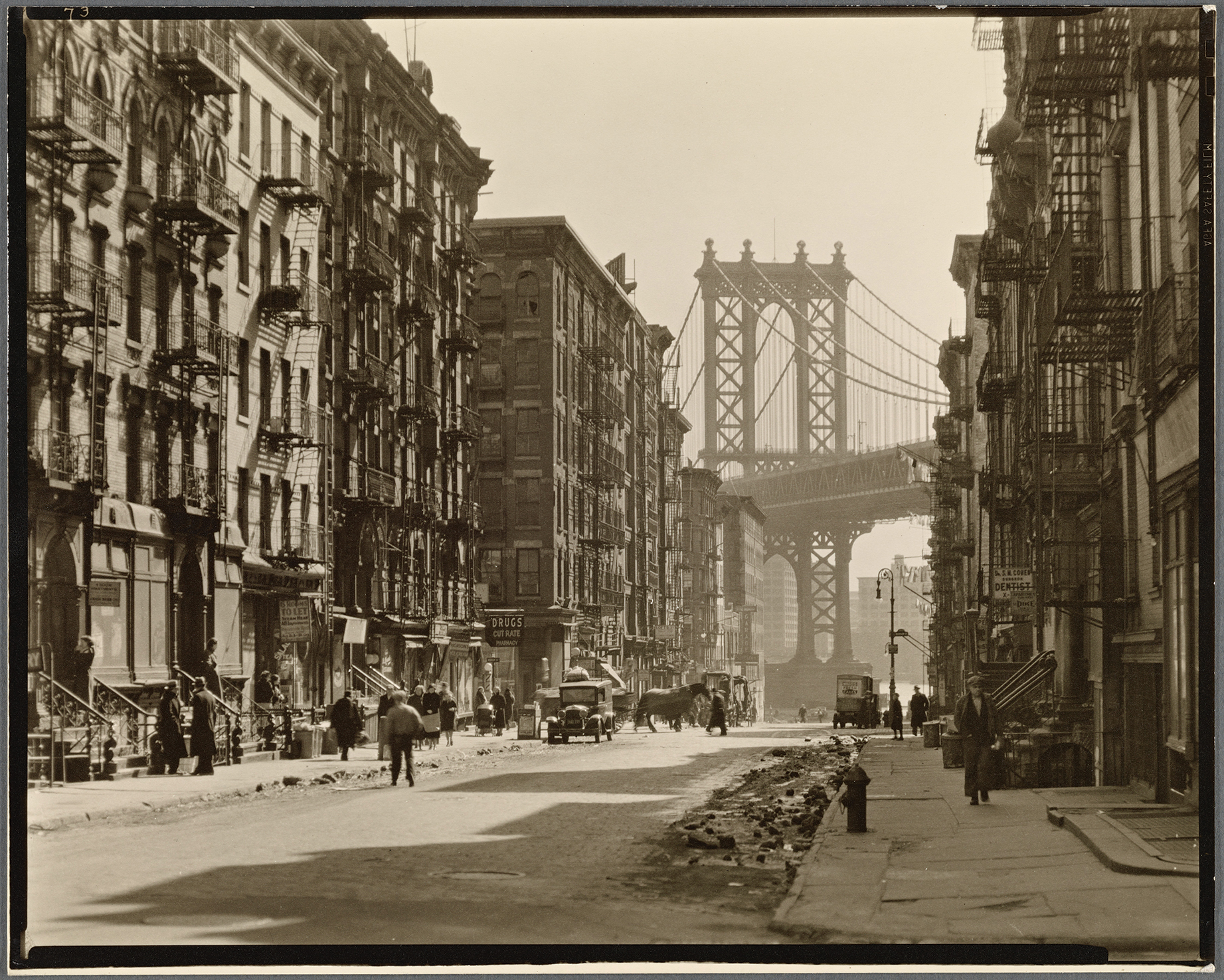 Vintage photo of a New York City street with the Manhattan Bridge in the background. Lined with tall buildings, people and cars bustle in a 1930s scene