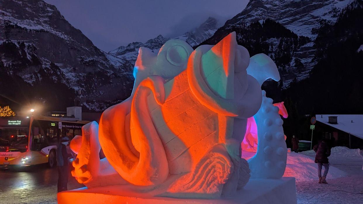 Red and pink lights on a snow sculpture of a sea monster and a ship with snow covered mountains and a bus in the background