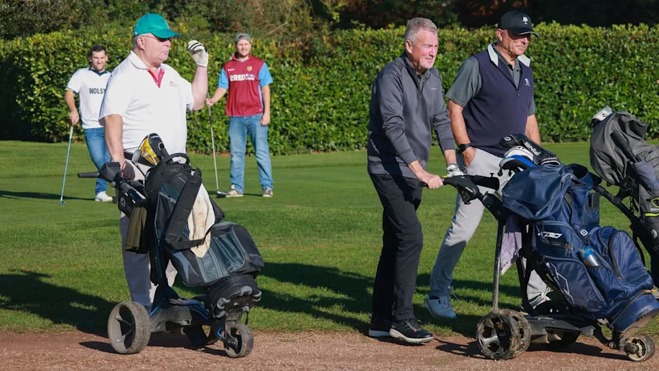  Three traditionally dressed golfers walk off the tee while two other golfer in football kits and jeans wait behind. 