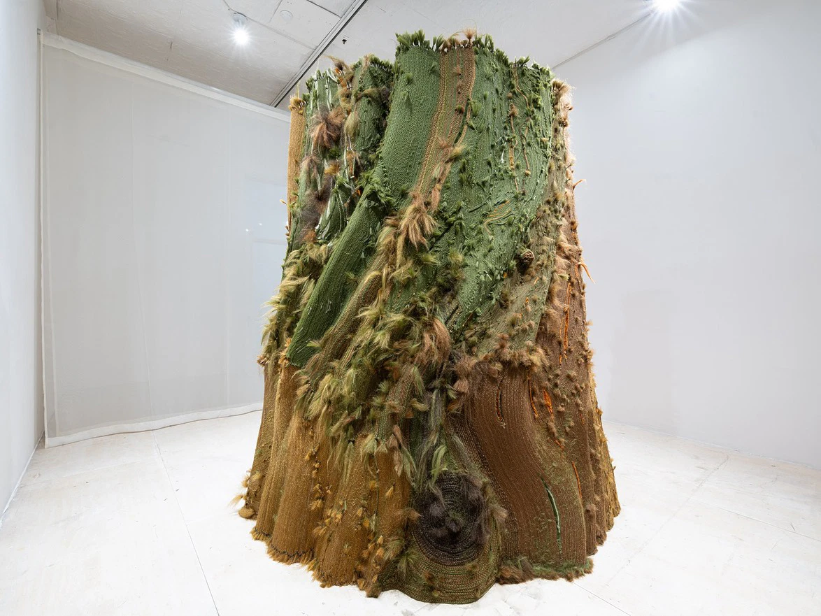 Large, textured abstract sculpture with green and brown hues displayed in a white gallery space with spotlights.