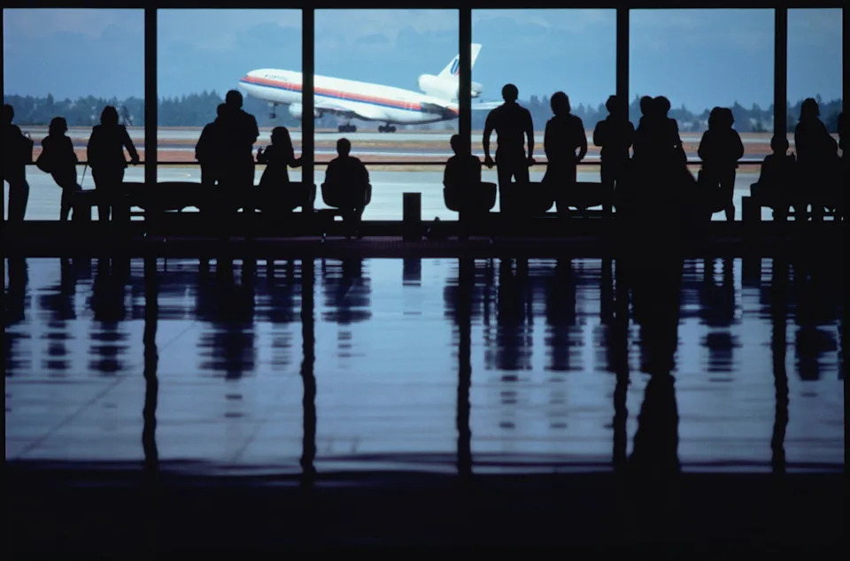 A group of people stands in an airport viewing area, watching a jet take off through the window.<p>Stock Photo via Getty Images</p>