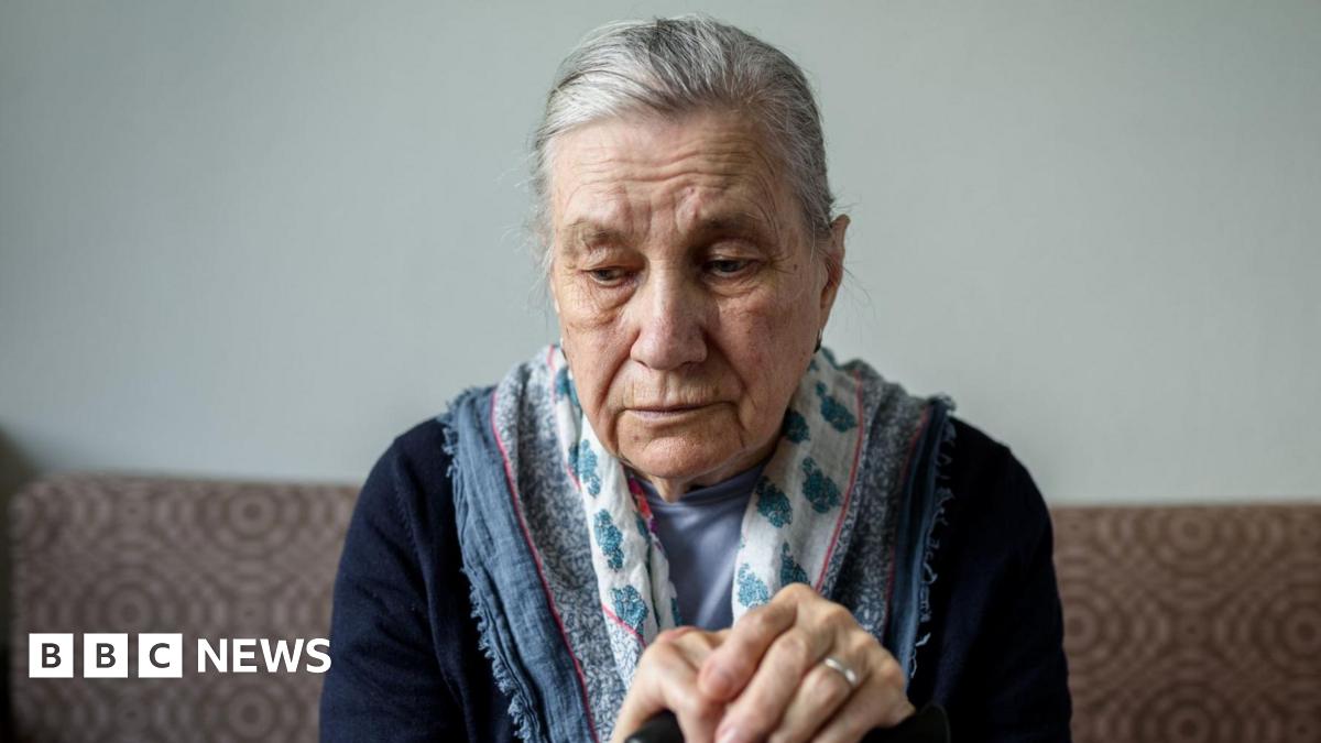 A woman with dark hair and a multiple colour jumper siting on a green sofa inside her living room.