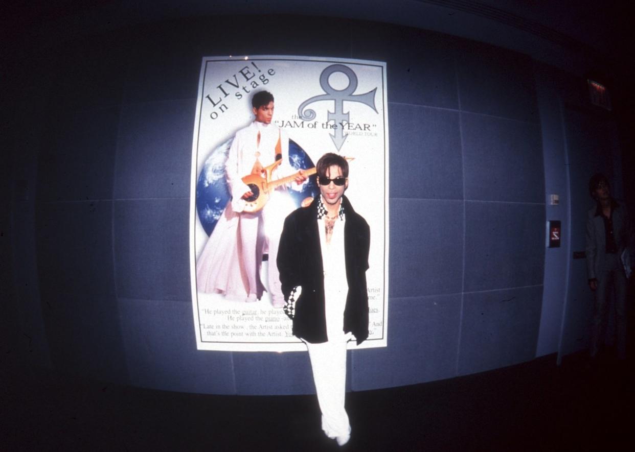 Prince posing near a poster promoting his live performance. - 