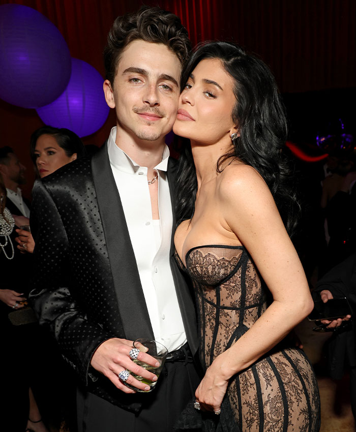 Timothée Chalamet at an event with a guest, dressed in a black tuxedo jacket and white shirt, posing for a photo.