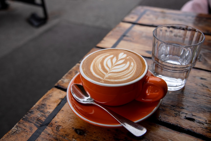 A coffee in a red cup with latte art on the foam, sitting atop a wooden table next to a glass of water
