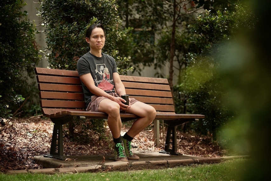 William, young, sits on a park bench, serious, wears shorts and black tee with a cartoon of a couple dancing in formal clothes.