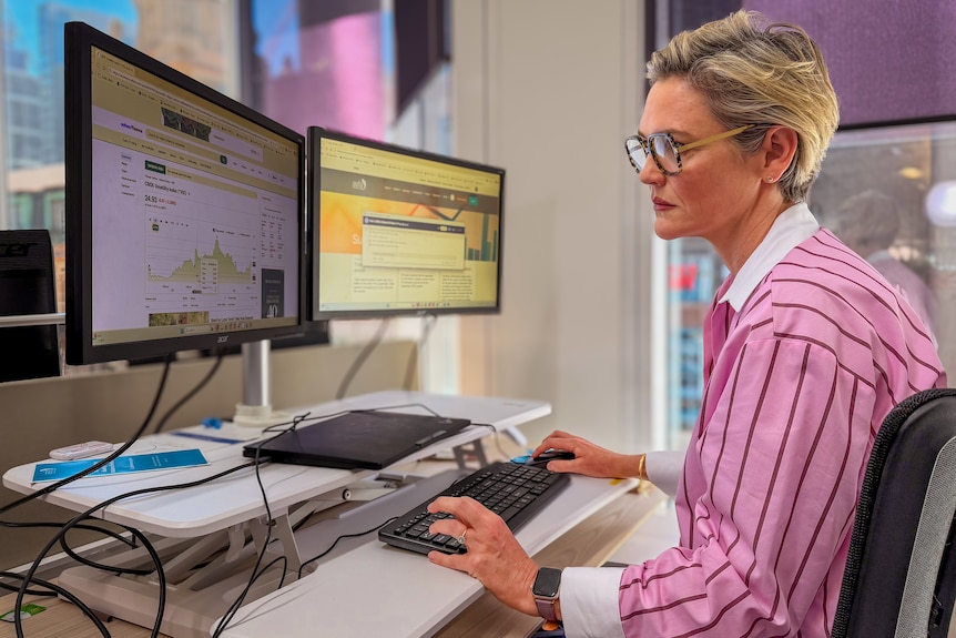A woman with short, blonde hair sits in front of computer screens at a desk in an office.