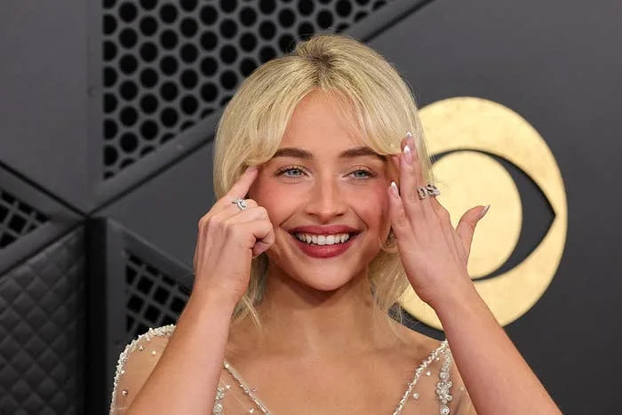 Person smiling on a red carpet with embellished dress, touching face with both hands, backdrop featuring geometric patterns and CBS logo