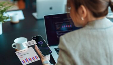 A woman with dark hair in a pony tail wearing a gray blazer looks at both a laptop and phone both displaying various bar charts.