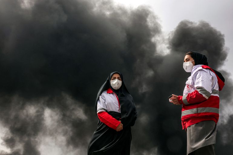 Women members of Iran's Red Crescent society stand near smoke plumes from an ongoing fire following an overnight airstrike on the Shahran oil refinery in northwestern Tehran on March 8, 2026.