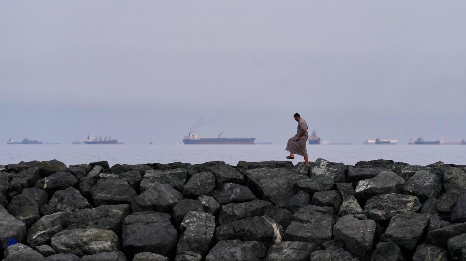 A man walks on rocks along the shore as oil tankers and cargo ships line up in the Strait of Hormuz, as seen from Khor Fakkan, United Arab Emirates, Wednesday, March 11, 2026. (AP Photo/Altaf Qadri)
