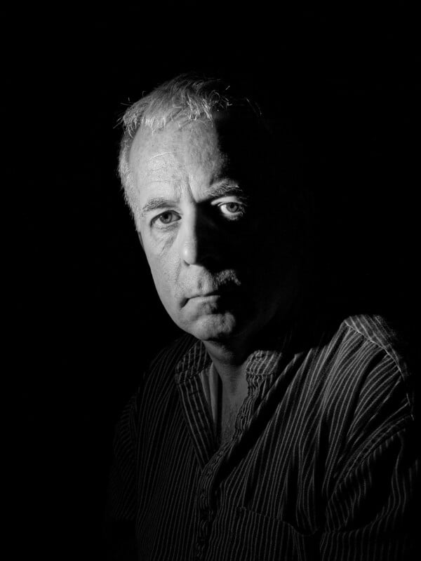 Black and white portrait of an older man with light hair and a serious expression, wearing a striped shirt, partially illuminated by dramatic lighting from the left, with the background in deep shadow.