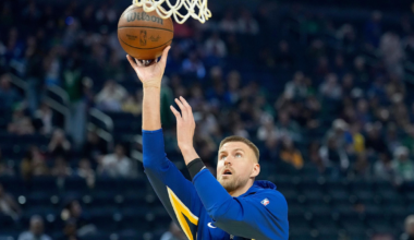 A basketball player in a blue and yellow warm-up jacket prepares to make a layup shot during a game with a crowd in the background.