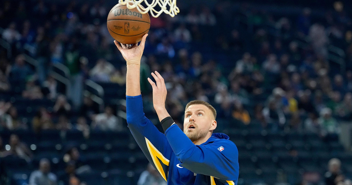 A basketball player in a blue and yellow warm-up jacket prepares to make a layup shot during a game with a crowd in the background.