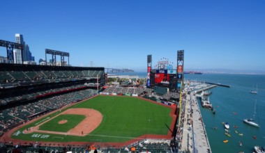 A sunny baseball stadium overlooks a green field with players, a dock with boats and kayaks, and a city skyline in the background under a clear blue sky.