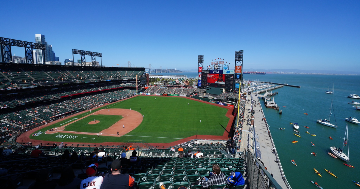 A sunny baseball stadium overlooks a green field with players, a dock with boats and kayaks, and a city skyline in the background under a clear blue sky.