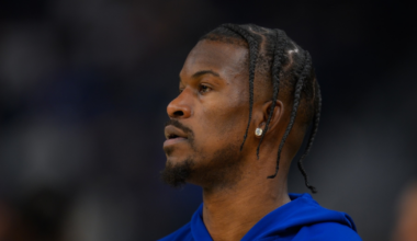 A man with braided hair and a small beard wears a blue hoodie and a diamond earring, looking intently to his left against a blurred background.