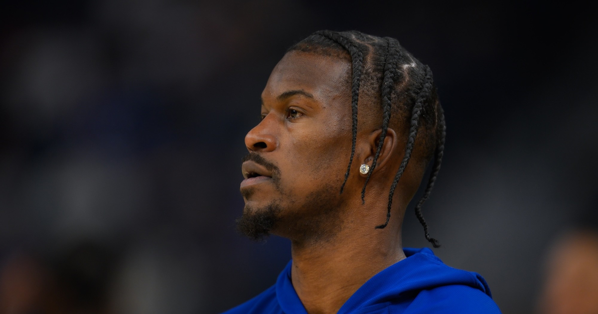 A man with braided hair and a small beard wears a blue hoodie and a diamond earring, looking intently to his left against a blurred background.