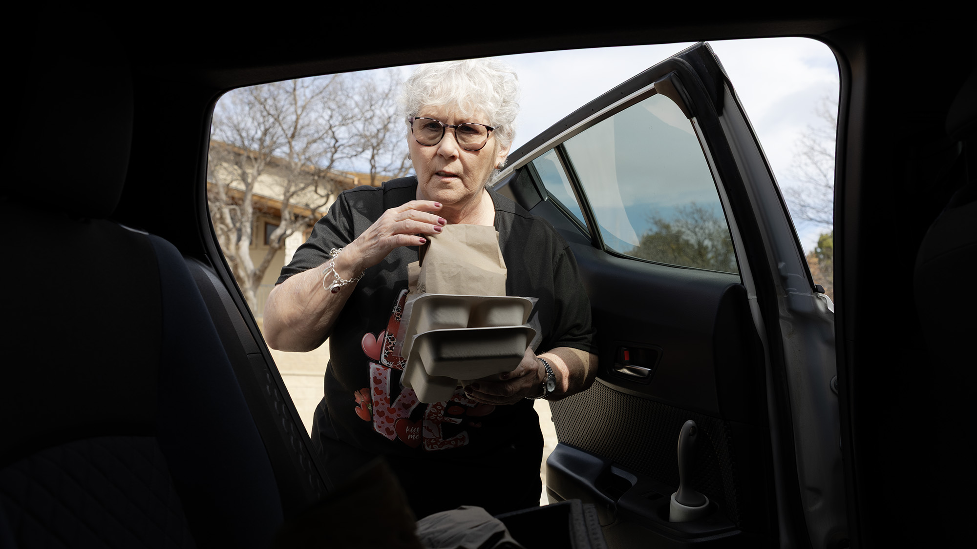 An older woman with short white hair and glasses picks up a stack of packaged meals from the back of a car.