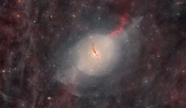 A starfield glows faintly with red glowing gas and dust.
In the center there is some sort of unusual galaxy with a red jet
emanating toward the upper right and a dark dust lane through
its center.  
Please see the explanation for more detailed information.