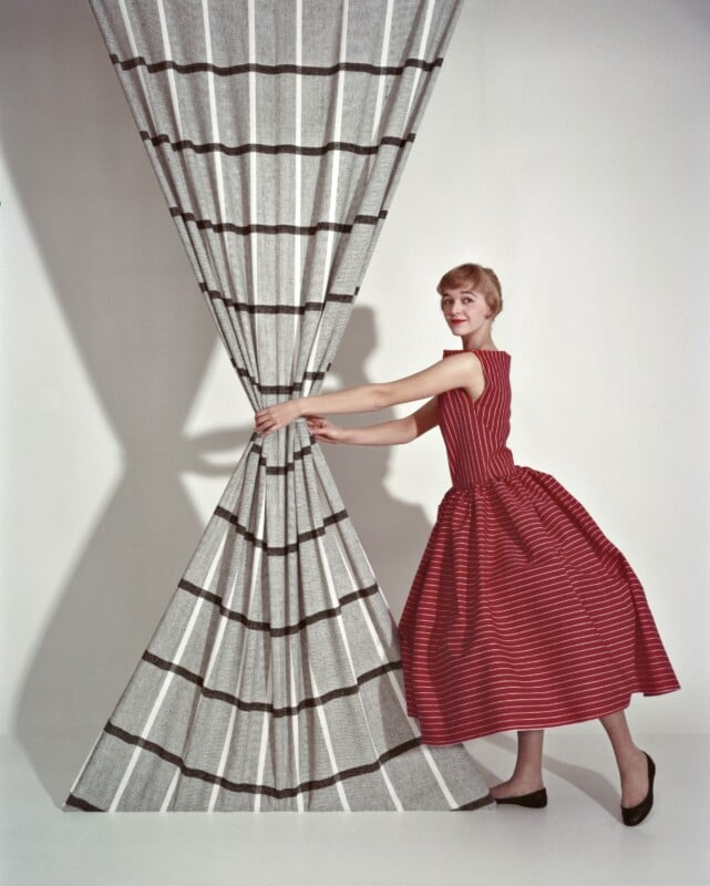 A woman in a red, sleeveless, striped dress pulls aside a large, gray curtain with black and white stripes, revealing an empty white background.